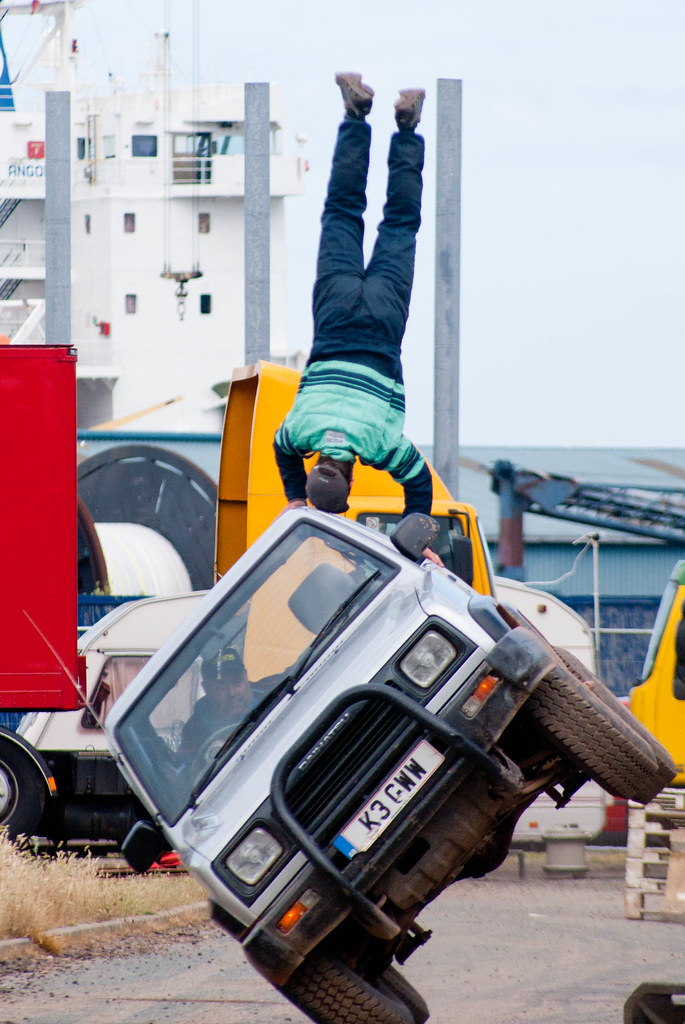 A truck is balanced on two wheels and a person is doing a handstand on the truck